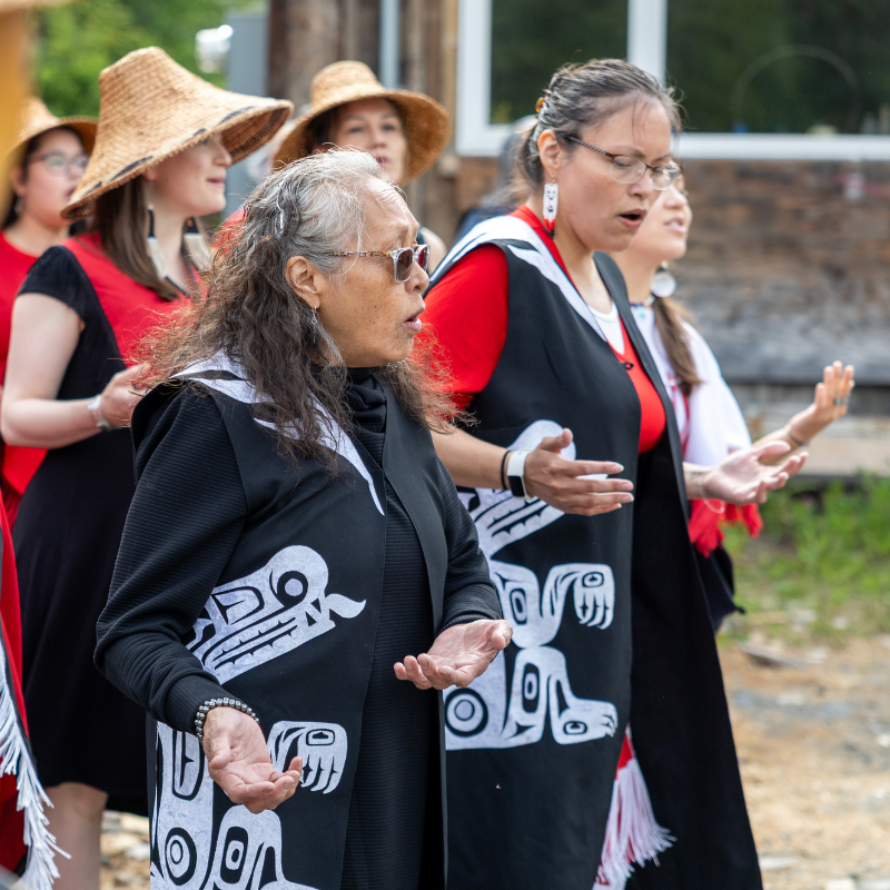 A group of women, wearing traditional clothing and hats, sing together outdoors.