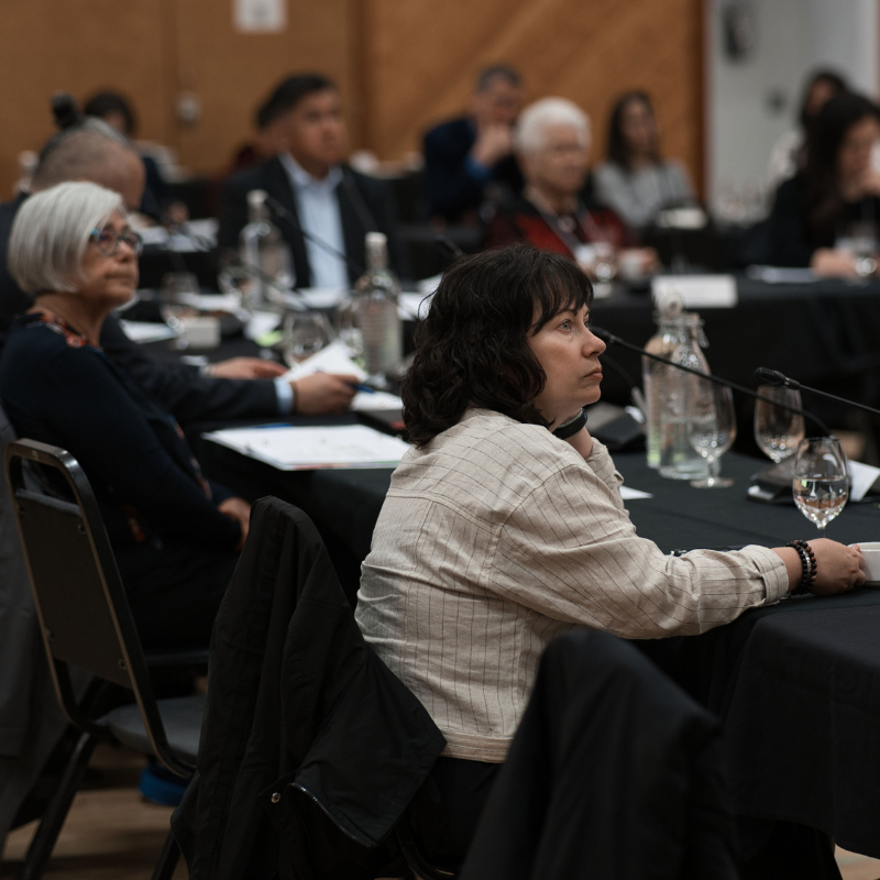 People sit attentively at a conference, focusing on a speaker. The setting is formal, with glasses and notebooks on tables.