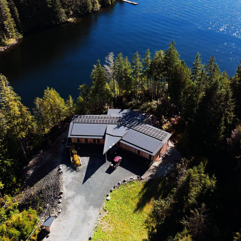 A wooden building, with solar panels on its black roof, stands at the end of a gravel driveway. It is surrounded by a lush, green forest and a sparkling, dark blue body of water.